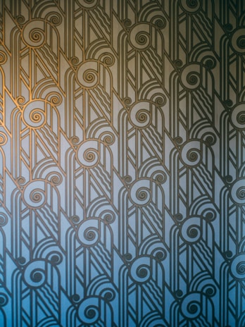 An Art Deco style pattern features intricate geometric shapes and swirling lines, arranged in a symmetrical and repetitive design. The pattern uses a gradient of blue and gold hues, creating a stylish and elegant visual effect.