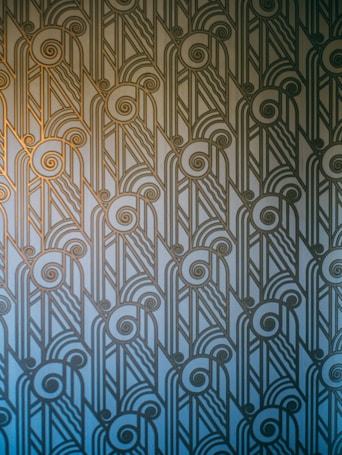 An Art Deco style pattern features intricate geometric shapes and swirling lines, arranged in a symmetrical and repetitive design. The pattern uses a gradient of blue and gold hues, creating a stylish and elegant visual effect.
