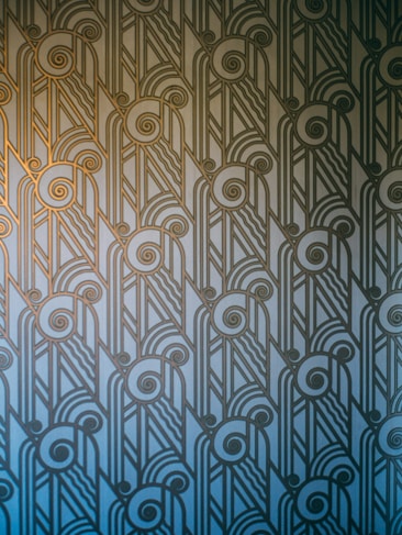 An Art Deco style pattern features intricate geometric shapes and swirling lines, arranged in a symmetrical and repetitive design. The pattern uses a gradient of blue and gold hues, creating a stylish and elegant visual effect.