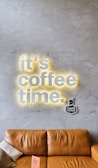 A stylish gray textured wall features the illuminated words 'it's coffee time' with a warm yellow glow. Below, a light brown leather couch is positioned with a small pillow and a pink booklet resting on one of its arms. A 'Café Cultura' logo is situated near the text on the wall.