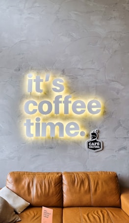 A stylish gray textured wall features the illuminated words 'it's coffee time' with a warm yellow glow. Below, a light brown leather couch is positioned with a small pillow and a pink booklet resting on one of its arms. A 'Caf&eacute; Cultura' logo is situated near the text on the wall.