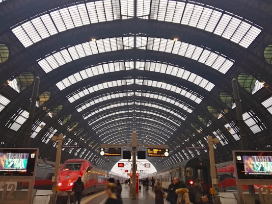 A large, arched train station platform featuring an intricate glass and steel roof. The scene includes two trains on either side of a central platform, with several people walking and digital displays showing information.