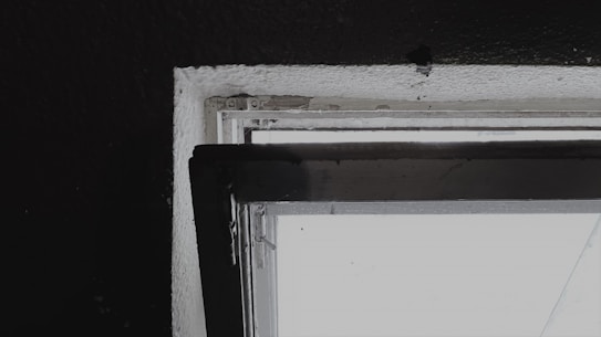 A close-up view of a window frame against a dark wall. The window appears to be slightly open with visible details of its structure, including the frame and hinges. The lighting creates a contrast between the bright window and the dark surrounding wall.