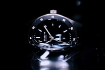 A close-up view of a sleek black wristwatch with a glossy dial and metallic case. The watch features luminous hands and hour markers, with minute indicators around the bezel. The lighting creates reflections and highlights on the watch surface, enhancing its luxurious appearance.