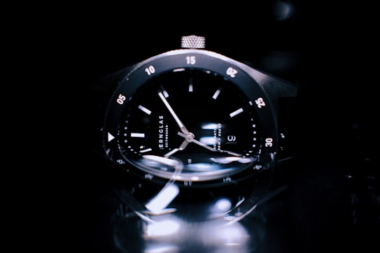 A close-up view of a sleek black wristwatch with a glossy dial and metallic case. The watch features luminous hands and hour markers, with minute indicators around the bezel. The lighting creates reflections and highlights on the watch surface, enhancing its luxurious appearance.