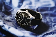 A sleek black wristwatch with a silver bezel rests on a textured, deep blue fabric. The watch face is dark with minimalistic hour markers and a date window, conveying a sense of elegance and modern style.