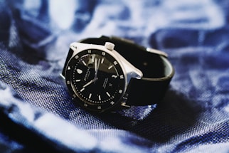 A sleek black wristwatch with a silver bezel rests on a textured, deep blue fabric. The watch face is dark with minimalistic hour markers and a date window, conveying a sense of elegance and modern style.