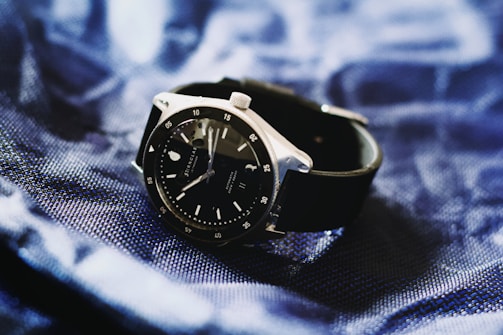 A sleek black wristwatch with a silver bezel rests on a textured, deep blue fabric. The watch face is dark with minimalistic hour markers and a date window, conveying a sense of elegance and modern style.