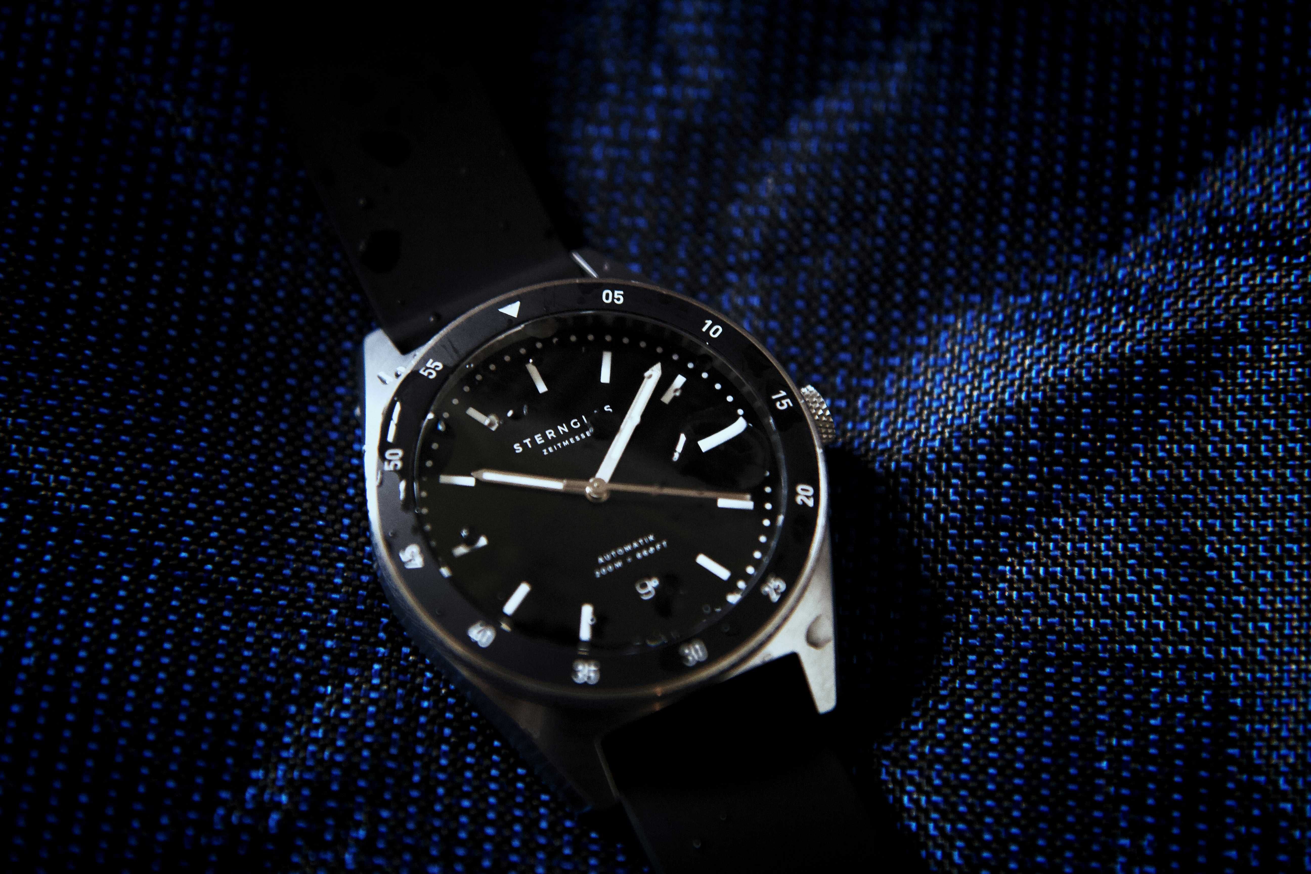 a black watch on a blue surface, The Sternglas Marus is a classic automatic diving watch with a rotating bezel and a water resistance of up to 20 ATM. https://www.sternglas.com/products/marus-black-rubber