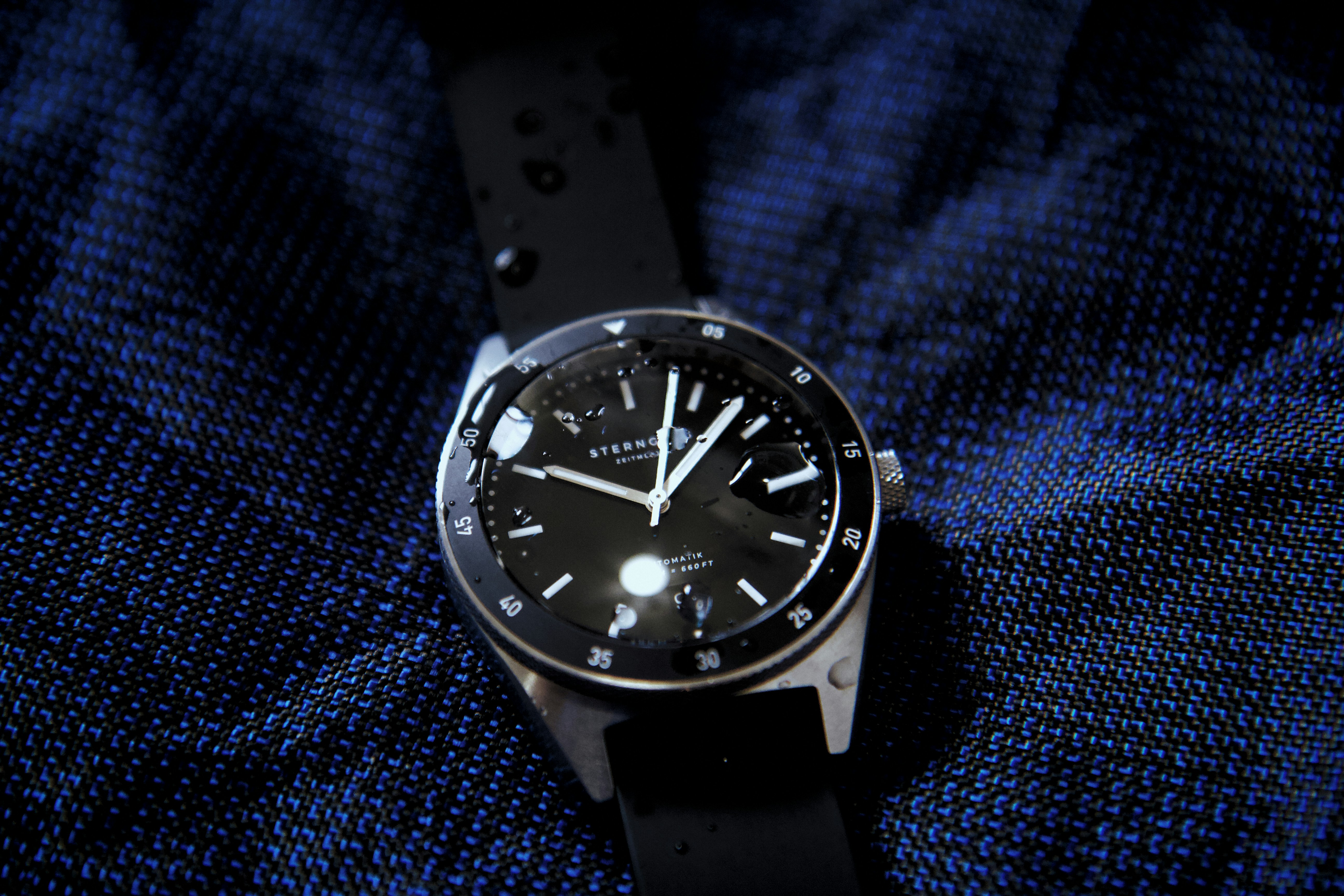 a black watch on a blue surface, The Sternglas Marus is a classic automatic diving watch with a rotating bezel and a water resistance of up to 20 ATM. https://www.sternglas.com/products/marus-black-rubber