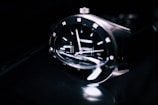 A close-up of a sleek and modern wristwatch with a reflective black face, silver hour markers, and polished metallic casing. The watch is shown at an angle, emphasizing its craftsmanship and the glossy texture of the materials.
