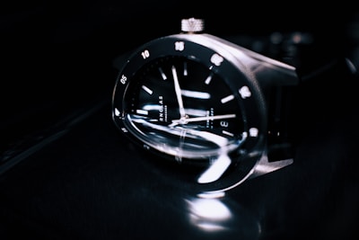 A close-up of a sleek and modern wristwatch with a reflective black face, silver hour markers, and polished metallic casing. The watch is shown at an angle, emphasizing its craftsmanship and the glossy texture of the materials.