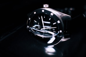 A close-up of a sleek and modern wristwatch with a reflective black face, silver hour markers, and polished metallic casing. The watch is shown at an angle, emphasizing its craftsmanship and the glossy texture of the materials.