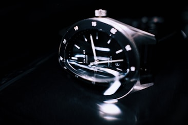 A close-up of a sleek and modern wristwatch with a reflective black face, silver hour markers, and polished metallic casing. The watch is shown at an angle, emphasizing its craftsmanship and the glossy texture of the materials.