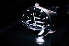 A close-up of a sleek and modern wristwatch with a reflective black face, silver hour markers, and polished metallic casing. The watch is shown at an angle, emphasizing its craftsmanship and the glossy texture of the materials.