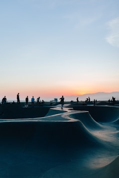 A skate park with curved ramps and silhouettes of people engaging in various activities under a sunset sky. The horizon is filled with soft orange and purple hues, creating a calming backdrop.