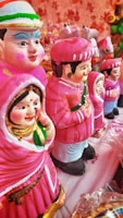A collection of colorful clay figurines depicting men and women in traditional attire. The figures are primarily dressed in pink with intricate facial features and decorative details. They are arranged in a line on a cloth-covered surface, surrounded by a soft, warm ambiance.