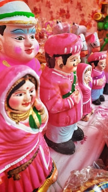 A collection of colorful clay figurines depicting men and women in traditional attire. The figures are primarily dressed in pink with intricate facial features and decorative details. They are arranged in a line on a cloth-covered surface, surrounded by a soft, warm ambiance.