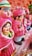 A collection of colorful clay figurines depicting men and women in traditional attire. The figures are primarily dressed in pink with intricate facial features and decorative details. They are arranged in a line on a cloth-covered surface, surrounded by a soft, warm ambiance.