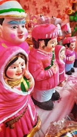 A collection of colorful clay figurines depicting men and women in traditional attire. The figures are primarily dressed in pink with intricate facial features and decorative details. They are arranged in a line on a cloth-covered surface, surrounded by a soft, warm ambiance.