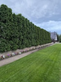 A neatly manicured green lawn stretches in the foreground, bordered by a path and a floral arrangement of colorful flowers. Behind the path, there is a dense line of tall, lush coniferous trees creating a natural fence. In the distance, a modern-looking house with a grey roof sits against a cloudy sky.