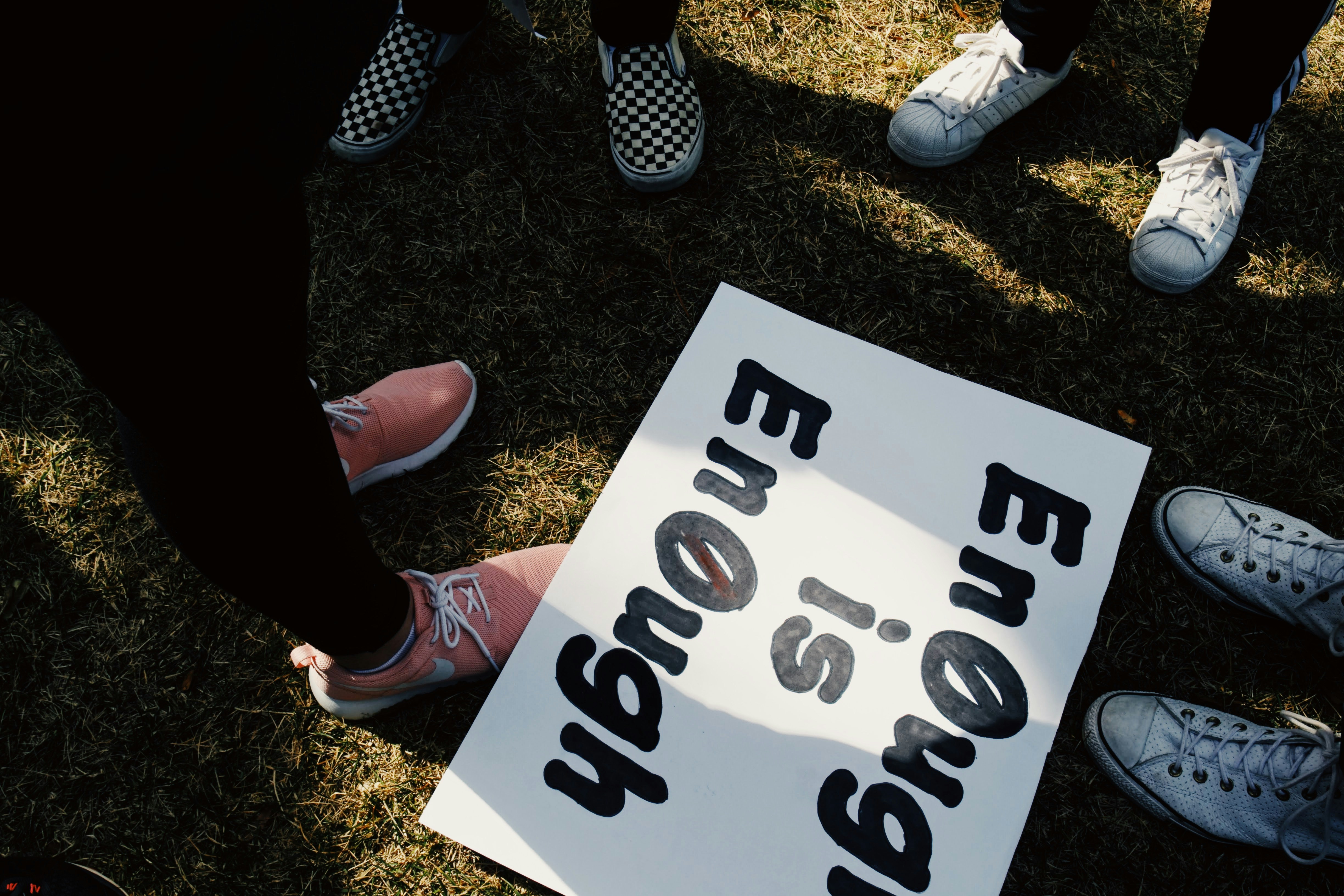 A white sign with large black text reading "ENOUGH IS ENOUGH" lies on grass, surrounded by various sneakers.