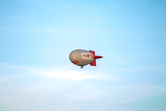 A large airship with a grey body and red tail floats against a clear blue sky. The airship features branding and text on its side.