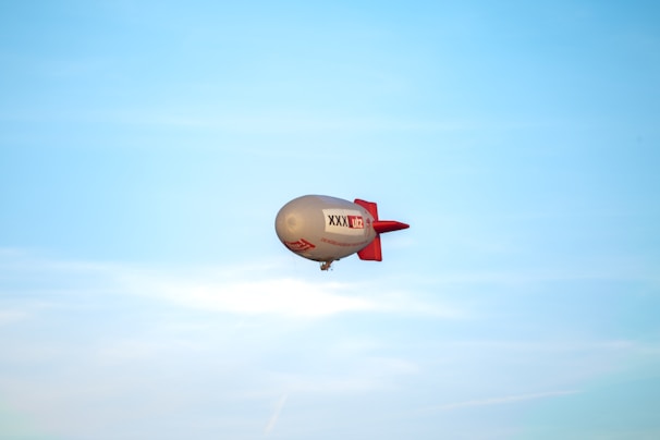 A large airship with a grey body and red tail floats against a clear blue sky. The airship features branding and text on its side.