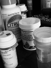 A collection of various medication bottles, including probiotics and fiber supplements, positioned on a surface. The bottles have prominent labels and are closely arranged together.