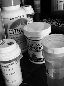 A collection of various medication bottles, including probiotics and fiber supplements, positioned on a surface. The bottles have prominent labels and are closely arranged together.