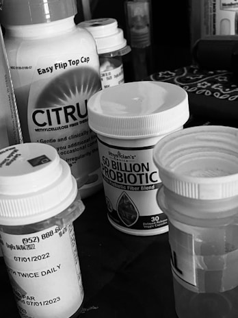 A collection of various medication bottles, including probiotics and fiber supplements, positioned on a surface. The bottles have prominent labels and are closely arranged together.