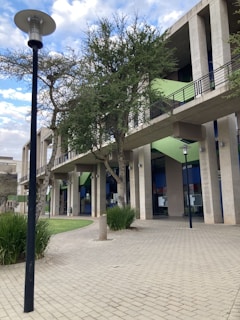 A modern architectural building with a minimalist design features large columns and an overhanging walkway. The structure integrates natural green elements with surrounding trees and landscaped grass. An outdoor setting with lamp posts enhancing the walkway's aesthetic. The backdrop contains a partially cloudy sky, adding depth to the scene.