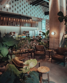 A cozy café interior with a modern, industrial style. The setting includes brown leather chairs and wooden tables surrounded by lush green plants. Exposed pipes and a decorative wall structure add to the ambiance. Baristas are seen behind a counter, with a warm light creating a relaxed atmosphere.
