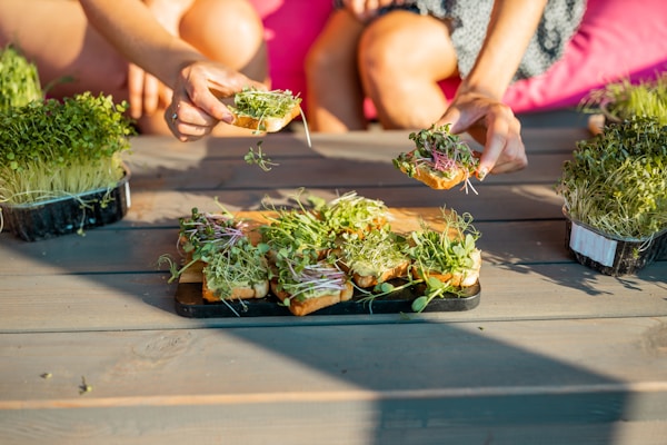 A group of small toasts topped with fresh green microgreens is placed on a wooden outdoor table. Two people are actively reaching out to take pieces of toast, surrounded by trays of more microgreens. The setting suggests a casual, alfresco dining experience with a focus on fresh, healthy ingredients.
