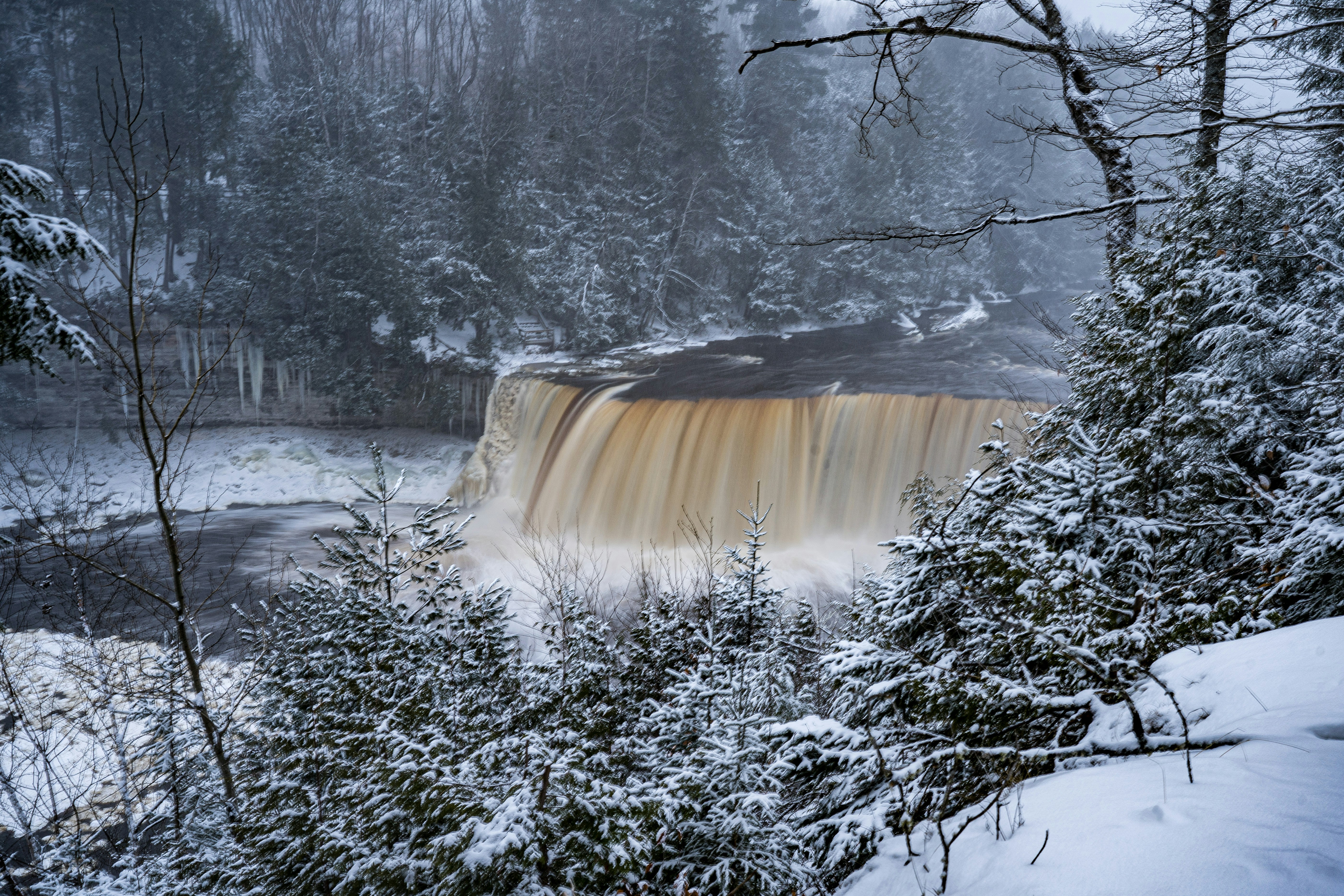 a snowy landscape with trees and a body of water with Tahquamenon Falls in the background