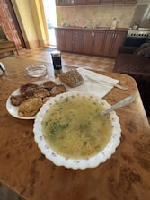 A kitchen setting with a table featuring a bowl of soup garnished with herbs, a plate of patties, and a piece of seeded bread. A pepper shaker is also present on the table. The background shows wooden cabinets, a countertop with kitchen items, and a stove, creating a cozy and homey atmosphere.