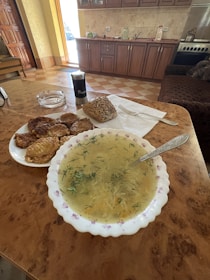 A kitchen setting with a table featuring a bowl of soup garnished with herbs, a plate of patties, and a piece of seeded bread. A pepper shaker is also present on the table. The background shows wooden cabinets, a countertop with kitchen items, and a stove, creating a cozy and homey atmosphere.
