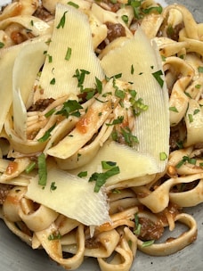 A delicious plate of pasta featuring ribbon-like noodles mixed with a savory meat sauce. The dish is garnished with thin slices of cheese and fresh green herbs, adding a touch of elegance and freshness.