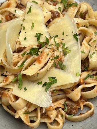 A delicious plate of pasta featuring ribbon-like noodles mixed with a savory meat sauce. The dish is garnished with thin slices of cheese and fresh green herbs, adding a touch of elegance and freshness.