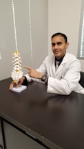 A person in a white coat, likely a medical professional, is seated at a desk and pointing to a spine model. The spine model is detailed with white vertebrae and yellow highlights. The setting appears to be an office or a clinical environment, with a neutral-colored wall and window nearby.