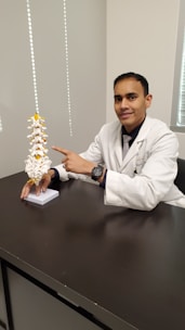 A person in a white coat, likely a medical professional, is seated at a desk and pointing to a spine model. The spine model is detailed with white vertebrae and yellow highlights. The setting appears to be an office or a clinical environment, with a neutral-colored wall and window nearby.
