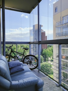A balcony with a cozy setup includes a blue couch with several pillows and a bicycle positioned nearby. The space is enclosed with glass panels, offering a clear view of neighboring apartment buildings and lush green trees under a bright blue sky.