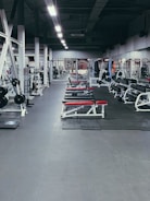 A spacious gym environment with various workout equipment neatly organized and aligned. The gym is empty, and there are several benches with red padding, weight racks, and gym machines. The floor is covered with rubber mats, and the lighting is bright, highlighting the equipment.