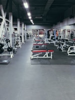A spacious gym environment with various workout equipment neatly organized and aligned. The gym is empty, and there are several benches with red padding, weight racks, and gym machines. The floor is covered with rubber mats, and the lighting is bright, highlighting the equipment.