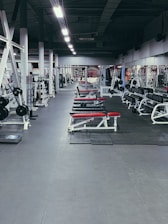 A spacious gym environment with various workout equipment neatly organized and aligned. The gym is empty, and there are several benches with red padding, weight racks, and gym machines. The floor is covered with rubber mats, and the lighting is bright, highlighting the equipment.