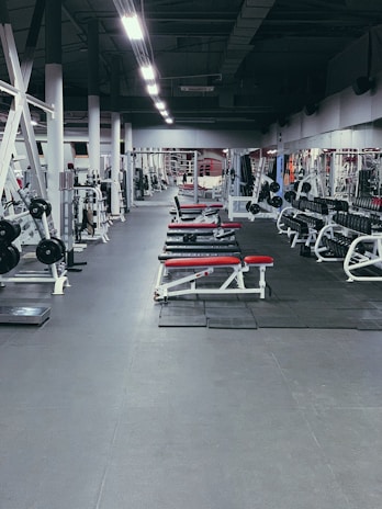 A spacious gym environment with various workout equipment neatly organized and aligned. The gym is empty, and there are several benches with red padding, weight racks, and gym machines. The floor is covered with rubber mats, and the lighting is bright, highlighting the equipment.