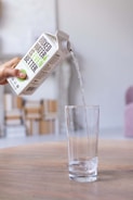 A hand is pouring water from a box into a tall clear glass. The box has the text 'Boxed Water Is Better' prominently displayed on it. The scene is set on a wooden table with a blurred background including some modern furniture.