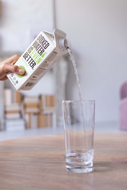 A hand is pouring water from a box into a tall clear glass. The box has the text 'Boxed Water Is Better' prominently displayed on it. The scene is set on a wooden table with a blurred background including some modern furniture.