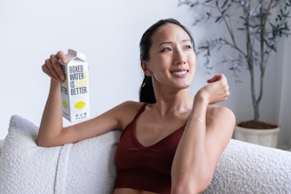 A person with a relaxed expression sits on a comfortable sofa, holding a carton of boxed water. The room appears bright and airy, with a potted plant in the background. The person is wearing a casual sleeveless top.