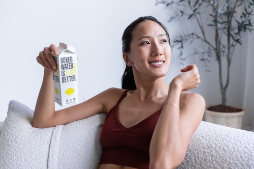 A person with a relaxed expression sits on a comfortable sofa, holding a carton of boxed water. The room appears bright and airy, with a potted plant in the background. The person is wearing a casual sleeveless top.